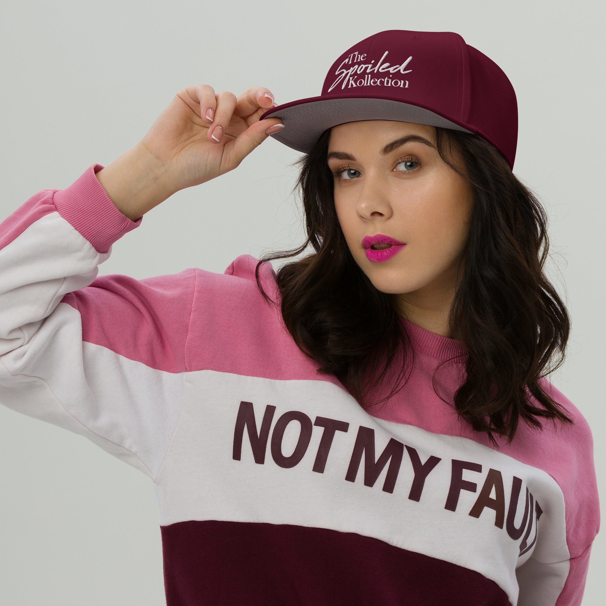 Model wearing a burgundy snapback hat and a pink sweatshirt with 'NOT MY FAULT' text, showcasing a trendy look.